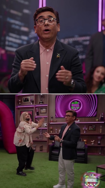 Um homem de terno e outro de peruca loira em um estúdio de televisão com grama artificial, prateleiras, troféus e telas de marca