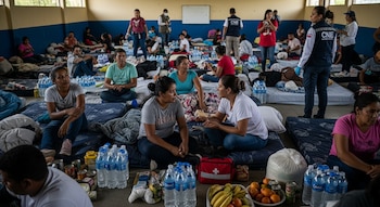 Personas en un albergue sentadas y acostadas en colchones. Botellas de agua, víveres y personal de la CNE con chalecos azules son visibles en la sala.