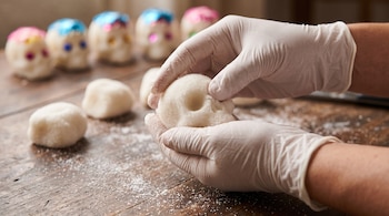 Dos manos con guantes blancos moldean una calavera de azúcar sobre una mesa de madera junto a porciones de masa y figuras decoradas.