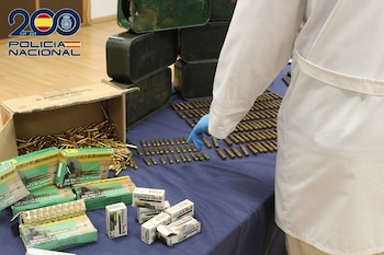 Una persona con guante azul señala munición sobre mesa azul. Hay cajas de cartón con proyectiles, envases de cartuchos y bidones verdes. Logo Policía Nacional