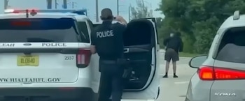 Un oficial de policía de pie junto a su patrulla apunta con un arma hacia una persona que permanece detenida en la calle