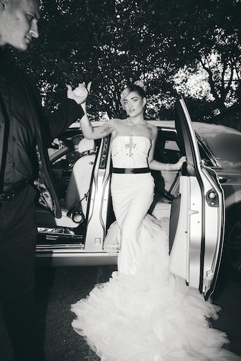 Foto em preto e branco de Marta Fort saindo de um carro de luxo, usando um vestido branco com cauda longa e um grande colar de cruz, segurando a mão de alguém