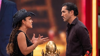 Una mujer con sombrero negro gesticula ante un hombre con camisa oscura, junto a un objeto dorado en forma de huevo sobre una corona.