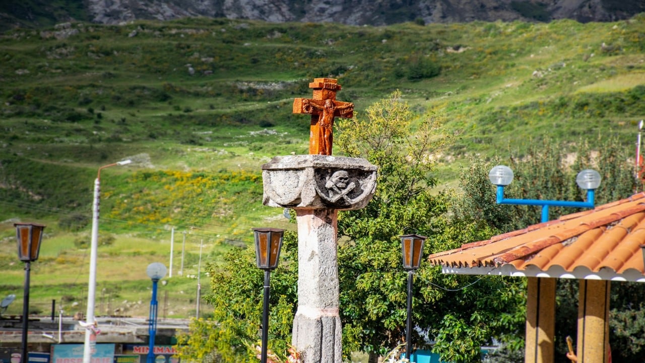 Una cruz de madera sobre un pedestal de piedra con relieves de calavera se ubica en un entorno natural con colinas verdes.
