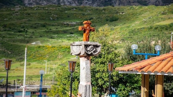Cruz de madera sobre pedestal de piedra con relieve de calavera. En el fondo, colinas verdes y árboles. Se ven farolas y un tejado de tejas de barro