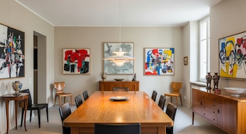 Vista de un comedor con una larga mesa de madera, sillas, una lámpara colgante, paredes beige con cuadros abstractos coloridos y aparadores de madera.