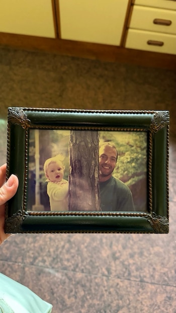 Una mano sostiene un portarretratos verde oscuro con una fotografía de un hombre sonriente y una niña junto al tronco de un árbol