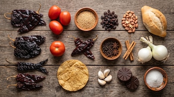 Chiles secos, jitomates, semillas, pasas, cacahuates, un bolillo, especias, cebollas, tortilla, ajos, tabletas de chocolate y azúcar sobre madera.