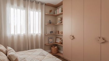 Un armario de tono beis junto a estanterías de rincón con juguetes y libros, al lado de una ventana con cortina blanca y una cama hecha.