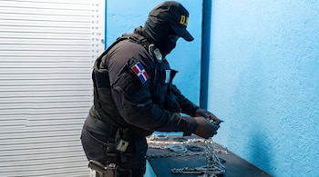 Persona con uniforme táctico negro y pasamontañas manipula esposas y cadenas sobre una mesa junto a una pared con escala de medición.