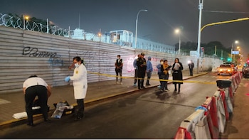 Escena nocturna en calle. Personal con batas blancas y policías investigan el suelo detrás de cinta amarilla. Un muro metálico con alambre de púas se ve al fondo