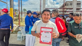Un hombre en primer plano con camiseta clara, sujeta un documento rojo de título de propiedad. Al fondo, personas y una valla metálica