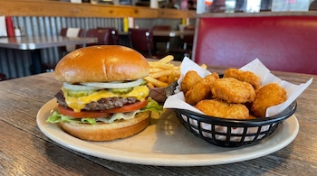 Una hamburguesa con queso, lechuga, tomate, cebolla y pepinillos en un plato, con un bol de nuggets y patatas fritas al lado, sobre una mesa de madera en un restaurante.