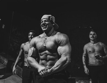 Hombre fisicoculturista musculoso con gorra y collar sonríe mientras flexiona sus brazos y abdomen. Otros dos hombres aparecen desenfocados detrás