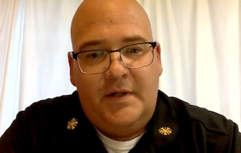 Un hombre calvo con gafas y uniforme oscuro, con insignias doradas en el cuello, mira al frente. Fondo de tela clara