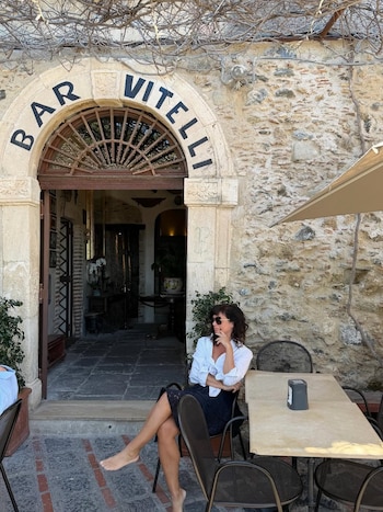 Griselda Siciliani está sentada descalça em uma mesa do lado de fora, em frente ao Bar Vitelli, um prédio de pedra com entrada em arco e telhado de videira.