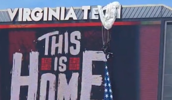 Paracaidista con paracaídas blanco y bandera de EE. UU. cuelga sobre una pantalla digital de Virginia Tech que muestra el texto 'THIS IS HOME'