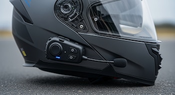 Primer plano de un casco de motocicleta negro mate con visor transparente y un dispositivo de intercomunicación con micrófono boom y luz azul encendida.