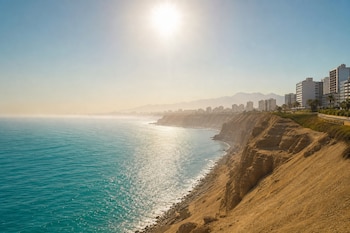Vista del mar de tono turquesa y acantilados de arena bajo la luz del sol brillante, con edificios en la costa al fondo.