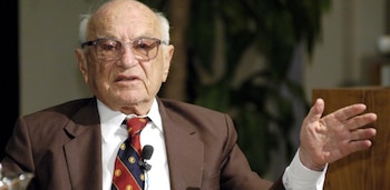 Milton Friedman viste traje marrón y corbata con micrófono de solapa, y gesticula con la mano abierta