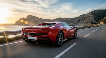 Vista trasera de un Ferrari deportivo rojo oscuro en movimiento por una carretera costera; el sol poniente ilumina el mar y las montañas rocosas al fondo.