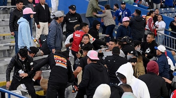 Un grupo de hombres con gorras y sudaderas forcejea entre las filas de asientos de un estadio con barandillas azules.