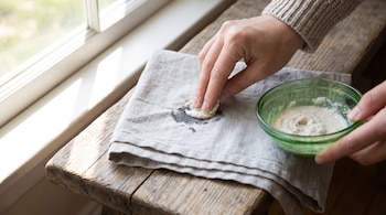 Mano de persona aplicando pasta blanquecina sobre mancha oscura en paño de lino gris. Cuenco verde con pasta sobre banco de madera junto a ventana.