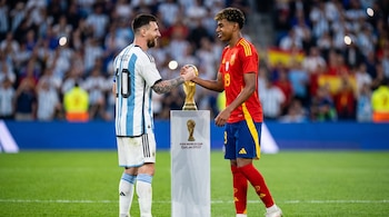 Lionel Messi y Lamine Yamal se estrechan las manos junto al trofeo de la Copa Mundial de la FIFA sobre un pedestal en un campo de fútbol con público.