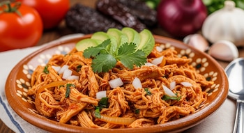 Plato de barro con tinga de pollo deshebrado, rodajas de aguacate, cilantro, cebolla blanca en cubos. Tomates, chiles secos y ajos al fondo.