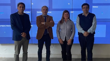 Cuatro personas (tres hombres y una mujer) de pie frente a una pared de pantallas azules con información y números en un entorno de oficina