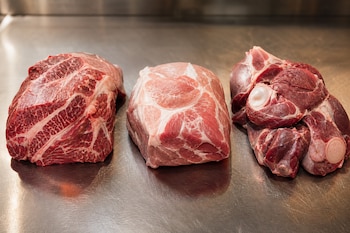 Tres cortes de carne cruda colocados en fila sobre una superficie de acero inoxidable.
