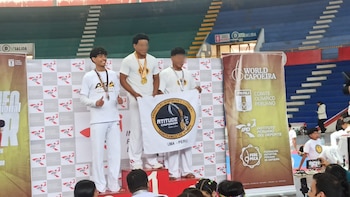 Tres jóvenes vestidos de blanco con medallas posan en un podio deportivo mientras dos de ellos sostienen una bandera