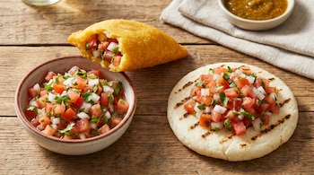 Vista aérea de una empanada mordida rellena de pico de gallo, una arepa con pico de gallo encima y un bol de la salsa, todo sobre madera.