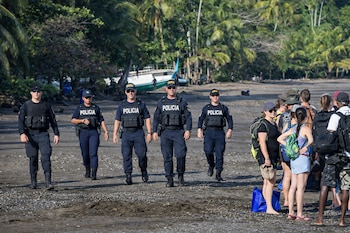 Cinco oficiales de policía con uniformes oscuros y chalecos tácticos caminan por una playa arenosa con palmeras. Un grupo de personas está a la derecha y hay botes al fondo.