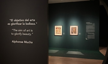 Una frase de Alphonse Mucha escrita en una pared oscura junto a la entrada a una sala con dos cuadros e información en pared verde