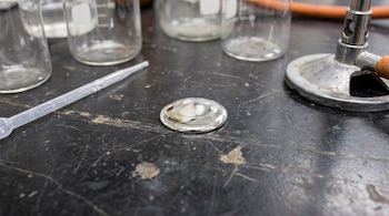 Una gota de mercurio líquido sobre una mesa de laboratorio junto a una pipeta de plástico, vasos de precipitado y un mechero Bunsen.