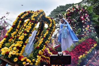 Estatua de la Virgen María y el Niño Jesús en una procesión, rodeada de arcos florales de rosas rojas y amarillas. Dos drones vuelan en el cielo