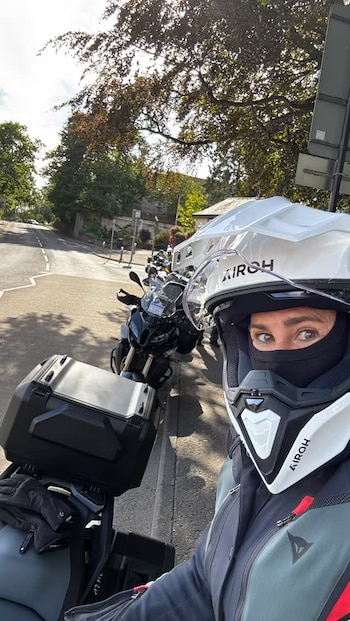 Una persona con casco blanco y pasamontañas posa en primer plano junto a motocicletas estacionadas en una calle