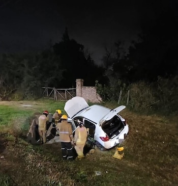 Vista nocturna de un auto blanco volcado en una zanja con el capó y el maletero abiertos, rodeado por cuatro rescatistas uniformados sobre el césped