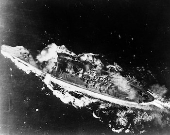 Vista aérea en blanco y negro de un acorazado clase Yamato gravemente dañado y humeante navegando en el océano, rodeado de estelas y explosiones