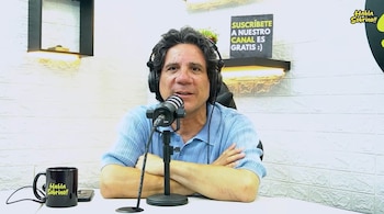Un hombre con auriculares y camisa celeste apoya los brazos sobre una mesa frente a un micrófono en un estudio con pared blanca