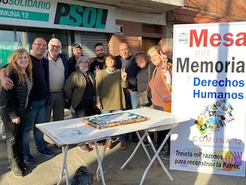 Grupo de personas posa sonriendo al aire libre. En primer plano, una mesa plegable blanca con una pieza de arte. A la derecha, un cartel grande blanco y rojo