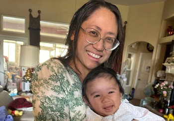 Una mujer con cabello oscuro y gafas sostiene a un bebé sonriente en sus brazos; ambos miran a la cámara y sonríen en un ambiente hogareño
