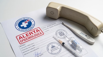 Documento de alerta epidemiológica junto a un auricular de teléfono, un termómetro digital, una jeringa y una ampolla de solución salina.