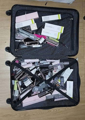 Una maleta negra abierta sobre un suelo de madera con decenas de cajas de productos cosméticos blancos, negros y rosados