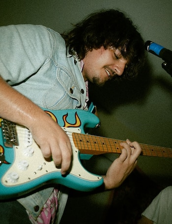 Hombre tocando guitarra eléctrica azul con diseño de llamas. Viste chaqueta de mezclilla. Manos en mástil y cuerdas. Se observa un micrófono