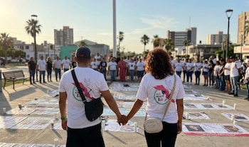 Un grupo de personas toma la mano de otra formando un círculo alrededor de carteles con fotos en el suelo de una plaza pública