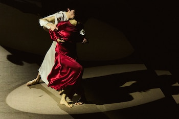 Dos personas descalzas vestidas con prendas de color rojo y blanco realizan un paso de baile dentro de un círculo de luz sobre el escenario