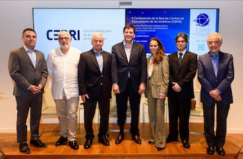 Siete personas, seis hombres y una mujer, de pie en un escenario de madera frente a dos pantallas grandes que muestran logotipos y texto de conferencia