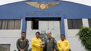 Cuatro hombres, dos con uniformes de aviador verdes y dos con camisas amarillas, posan frente a un edificio azul con la inscripción Grupo Aéreo 3